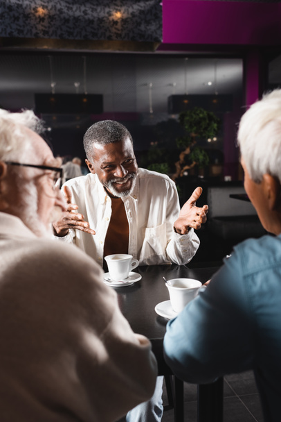 homem americano africano alegre apontando com as mãos enquanto conversa com amigos seniores desfocados no bar - Foto, Imagem