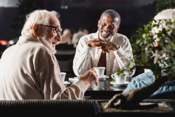старший чоловік в окулярах розмовляє з міжрасовими друзями, проводячи час у кафе
 - Фото, зображення
