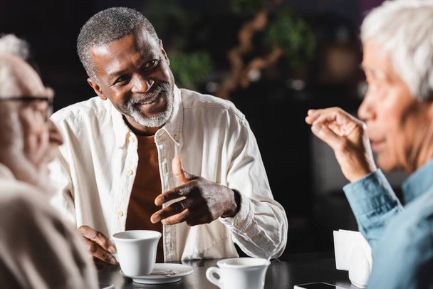 radosny Afrykanin amerykański człowiek gestykulując podczas rozmowy z zamazanymi starszymi przyjaciółmi w restauracji - Zdjęcie, obraz