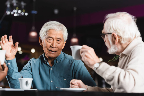 vanhempi aasialainen mies elehtii juodessaan kahvia ja puhuessaan monikansallisten ystävien kanssa - Valokuva, kuva