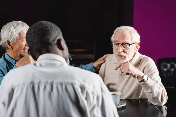 старший чоловік в окулярах вказує пальцем під час розмови з міжрасовими друзями в кафе
 - Фото, зображення