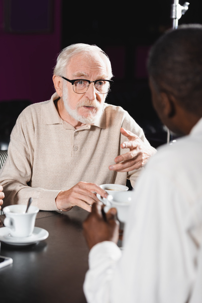 senior man in eyeglasses gesturing while talking to blurred african american man in cafe - Photo, Image