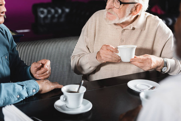 Ausgeschnittene Ansicht eines älteren Mannes mit Brille, der eine Kaffeetasse hält, während er Freund im Café zuhört - Foto, Bild