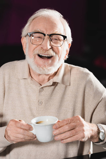 cheerful senior man in eyeglasses holding cup of coffee while laughing at camera - Photo, Image