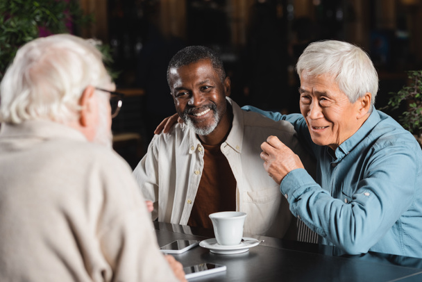 smiling asian man embracing shoulders of african american friend in cafe - Photo, Image