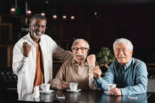 senior multicultural friends looking at camera and showing win gesture in cafe - Fotó, kép