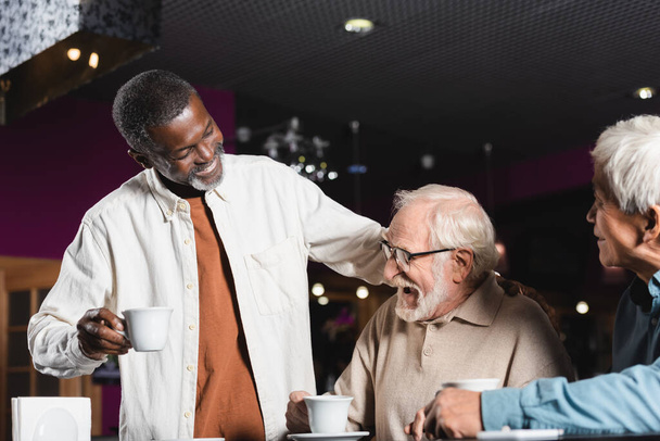 glimlachende Afrikaan amerikaanse man met koffie beker staan in de buurt van senior interraciale vrienden lachen in cafe - Foto, afbeelding