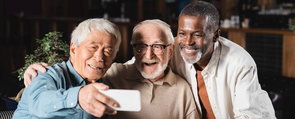 sonriente asiático hombre tomando selfie en smartphone con senior multiétnico amigos, bandera - Foto, imagen
