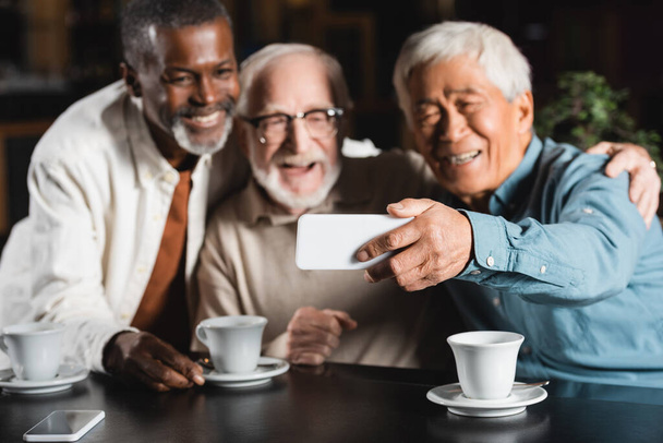 joyful multiethnic friends taking selfie on mobile phone in cafe, blurred background - Фото, изображение