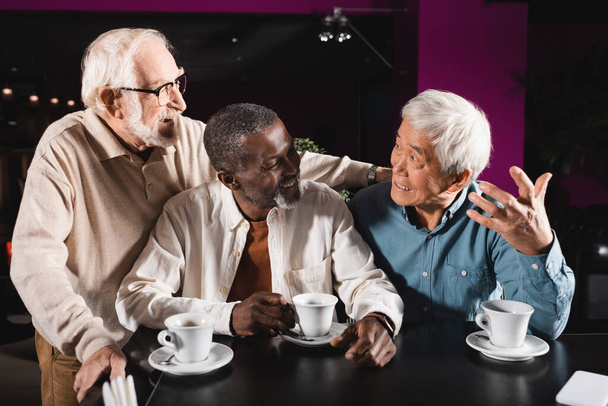 emocional asiático homem gestos enquanto falando com alegre multicultural amigos no café - Foto, Imagem