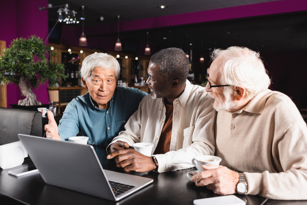 senior interracial amis pointant vers ordinateur portable tout en parlant dans le café - Photo, image