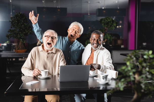 cheerful senior multiethnic friends looking at camera near laptop in cafe - Photo, Image