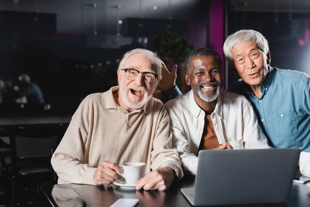 amigos multiétnicos seniores alegres que sorriem para a câmera perto do computador portátil no café - Foto, Imagem