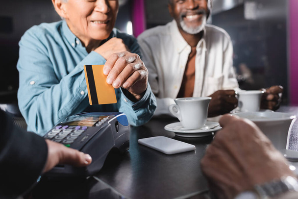 widok upraw seniora z karty kredytowej w pobliżu terminalu płatniczego i Afrykański amerykański człowiek w kawiarni - Zdjęcie, obraz