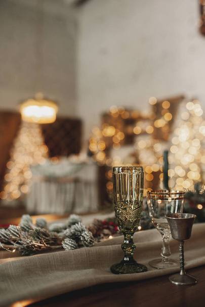 elegáns karácsonyi asztal beállítás bézs és fa színekben, üvegáru arany evőeszközökkel, gyertyák, pamut és koszorúk ellen bokeh háttér és karácsonyi belső, szelektív fókusz - Fotó, kép