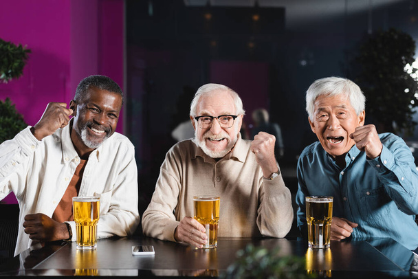 happy senior multiethnic friends showing win gesture while watching football championship in pub - Foto, Bild