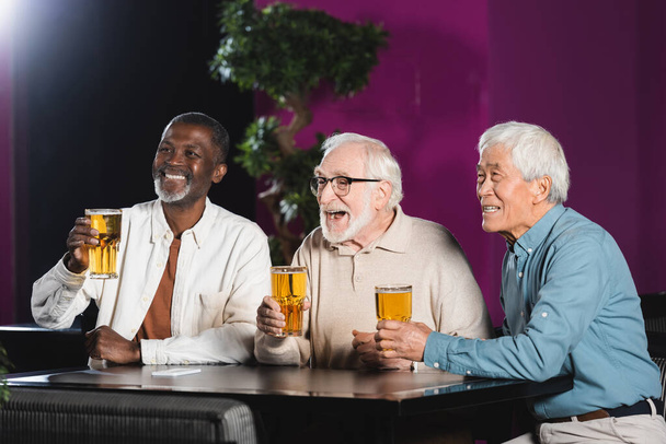 joyful elderly multiethnic friends with glasses of beer watching football match in pub - Photo, Image