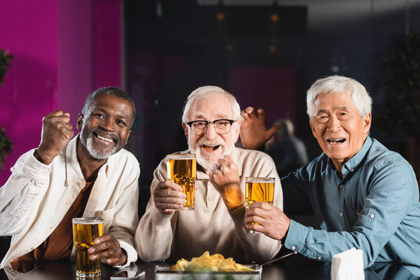 joyful senior multicultural friends with glasses of beer showing win gesture while watching football game in pub - Фото, зображення