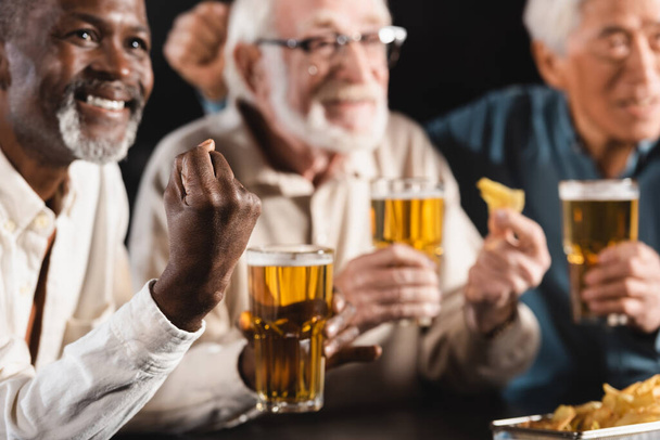 smiling african american man showing success gesture near blurred senior interracial friends watching football in pub - Photo, Image
