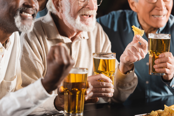 ostříhaný pohled na starší multietnické přátele, jak jedí chipsy a pijí pivo v hospodě - Fotografie, Obrázek