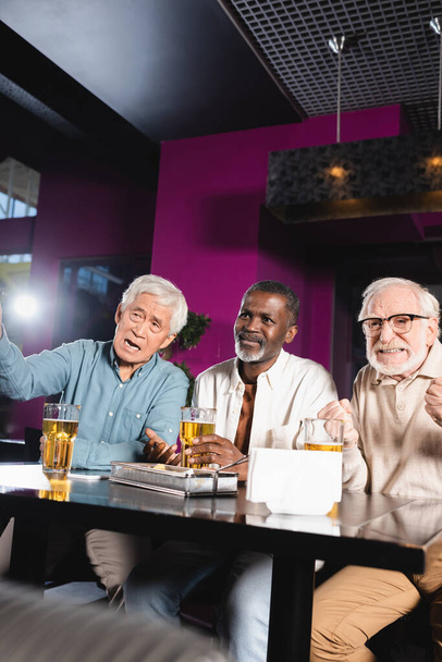 tenso hombre mayor con los puños cerrados viendo el partido de fútbol con amigos multiétnicos en el pub - Foto, imagen
