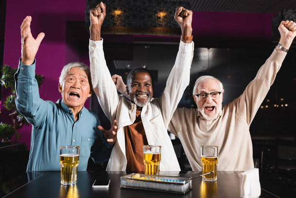 excited senior multiethnic friends watching football match in pub and showing win gesture - Foto, afbeelding