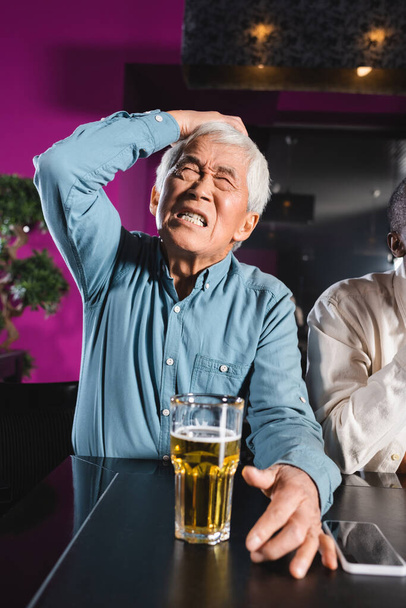 sorprendido sénior asiático hombre con cerrado ojos tocando cabeza mientras sentado con africano americano amigo en deportivo bar - Foto, imagen
