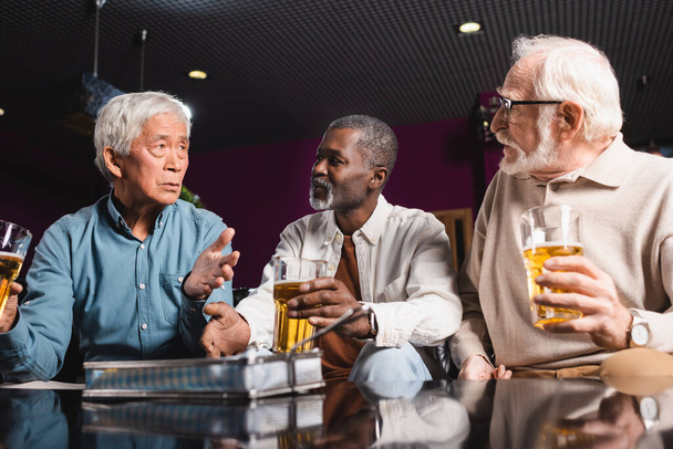 senior friends gesturing and holding glasses of beer during conversation in pub - Foto, immagini