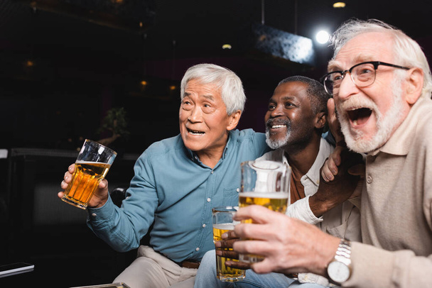 amazed senior man with open mouth watching football in beer pub with happy interracial friends - Foto, afbeelding