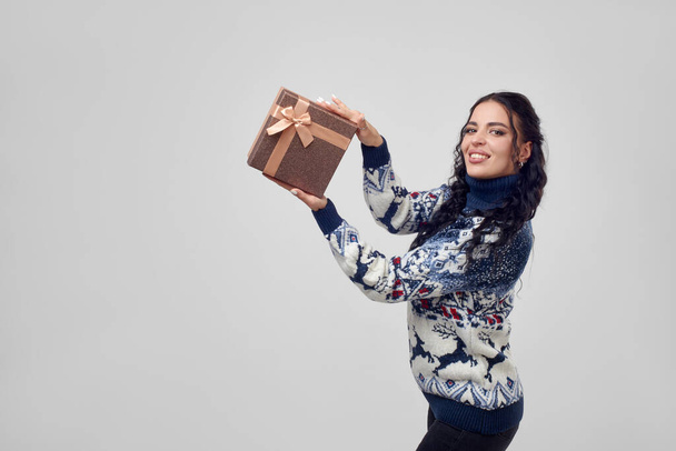 Жінка в різдвяному светрі демонструє подарункову коробку на сірому фоні
. - Фото, зображення
