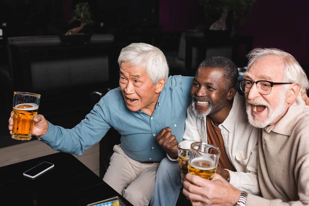 homme afro-américain souriant avec poing serré montrant match de football avec des amis interraciaux excités dans un pub - Photo, image
