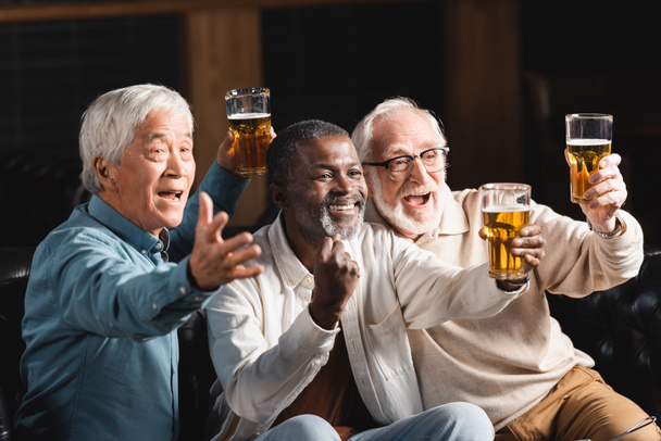hombre afroamericano senior mostrando gesto de triunfo cerca de amigos multiétnicos emocionados viendo fútbol en bar deportivo - Foto, imagen