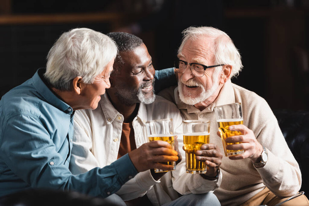 happy, elderly multiethnic friends holding glasses of beer while spending time in pub - Foto, imagen