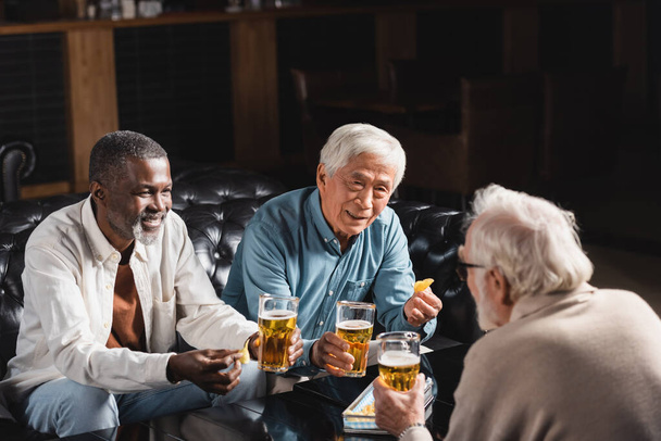 senior multicultural friends holding glasses of beer while spending time in pub - Foto, afbeelding
