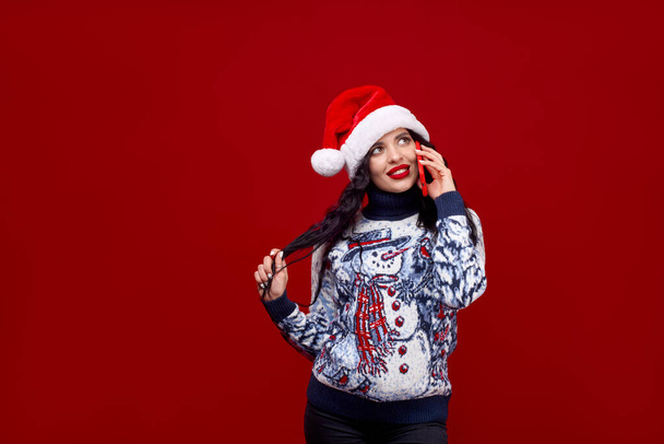 Świąteczne pozdrowienia przez telefon. Kobieta w noworocznej czapce i swetrze emocjonalnie rozmawia przez telefon na czerwonym tle i trzyma się za włosy.. - Zdjęcie, obraz