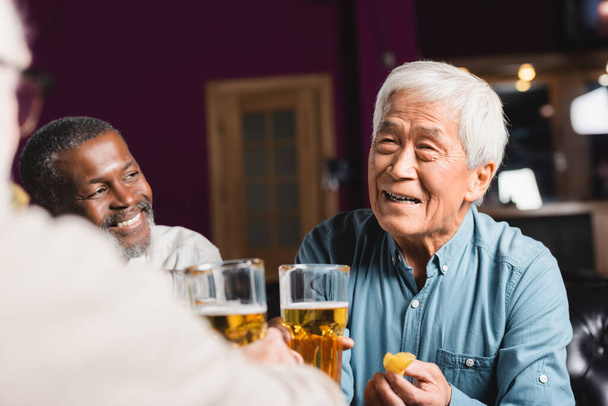 gelukkig aziatische man met bier en chips in de buurt wazig multi-etnische vrienden in pub - Foto, afbeelding