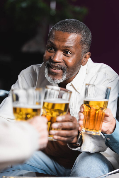senior african american man smiling while clinking blurred beer glasses with friends - Photo, Image