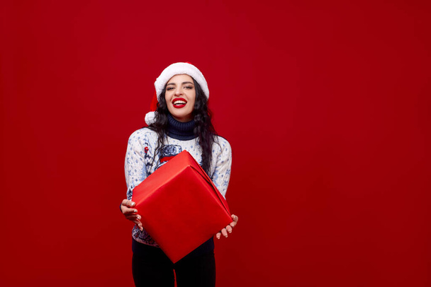 A young girl in a New Year's cap and sweater holds out a red box on a red background smiling. - Photo, Image