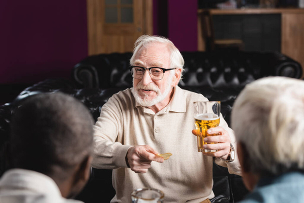 vidám idős férfi kezében chips és sör, miközben beszél homályos fajok közötti barátok kocsmában - Fotó, kép