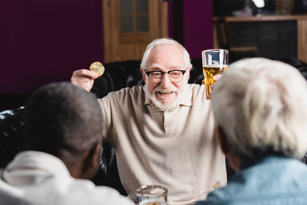 ενθουσιασμένος ηλικιωμένος άντρας με μπύρα και πατατάκια που μιλάει σε θολή πολυεθνικούς φίλους στην παμπ - Φωτογραφία, εικόνα