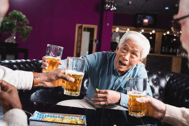 podekscytowany starszy Azji mężczyzna toasting z piwa w pobliżu rozmazane wielonarodowych przyjaciół w pubie - Zdjęcie, obraz