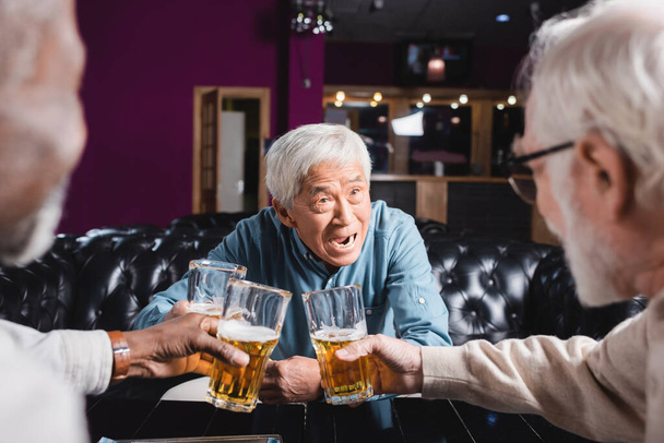 astonished asian man clinking glasses of beer with blurred interracial friends in pub - Fotó, kép