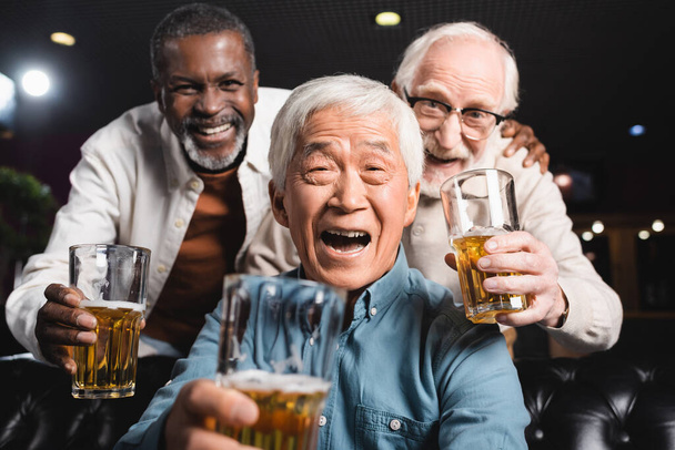 wesoły wieloetniczny przyjaciele trzymając okulary z piwem patrząc na aparat fotograficzny w pubie - Zdjęcie, obraz
