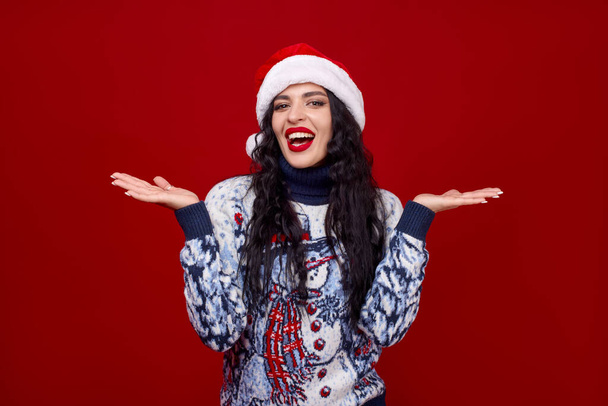 Uśmiechnięta młoda kobieta w świątecznej czapce na czerwonym tle podnosi ręce i patrzy w kamerę. Studio Sylwester sesji zdjęciowej. - Zdjęcie, obraz