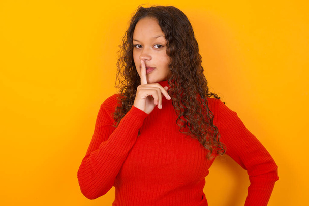  woman wearing red sweater standing against yellow background makes hush gesture, asks be quiet. Don't tell my secret or not speak too loud, please! - Photo, Image