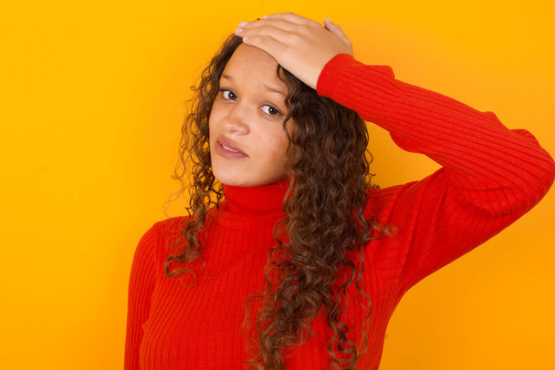 Oops, what did I do?  woman wearing red sweater standing against yellow background holding hand on forehead with frightened and regret expression. - Photo, Image
