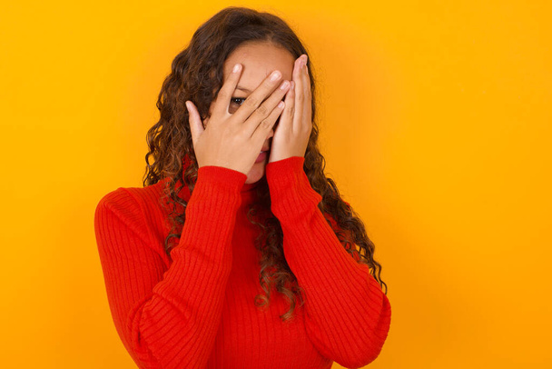  woman wearing red sweater standing against yellow background covering face with hands and peering out with one eye between fingers. Scared from something or someone. - Foto, Imagem