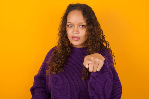カメラのインデックスフィンガー付きの黄色の背景ポイントに立った紫色のセーターを着用したショックを受けた女性. サプライズと広告コンセプト. - 写真・画像