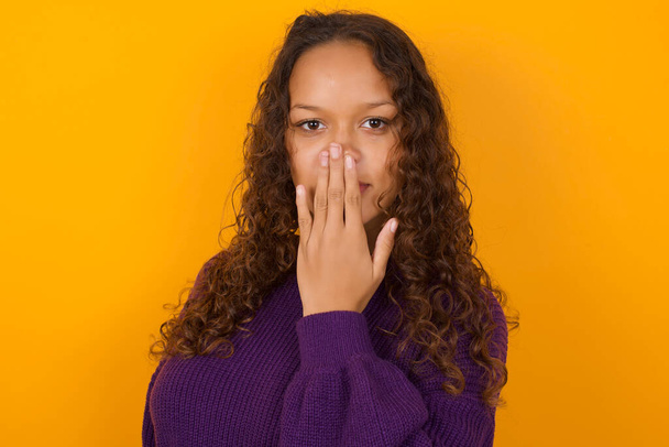 Oh! I think I said it! Close up portrait  woman wearing purple sweater standing against yellow background cover open mouth by hand palm, look at camera with big eyes. - Photo, Image