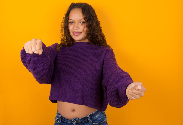 Portrait of charming woman wearing purple sweater standing against yellow background smiling broadly while holding hands over her head.  Confidence and relax concept. - 写真・画像